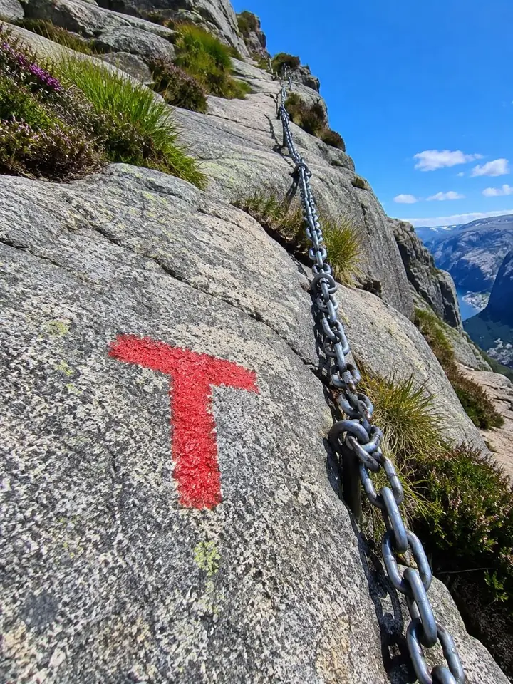 Ein rotes T-Wanderwegzeichen, das auf Granit neben einer festen Stahlkette am Kjerag-Weg aufgemalt ist